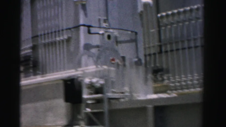 1957 Nevada 8mm frame showing railings, pipework, and service equipment along a concrete industrial exterior.