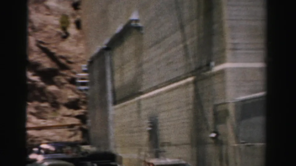 1957 Nevada 8mm frame with a concrete wall, metal railing, pipework, and adjacent rocky cliff face.