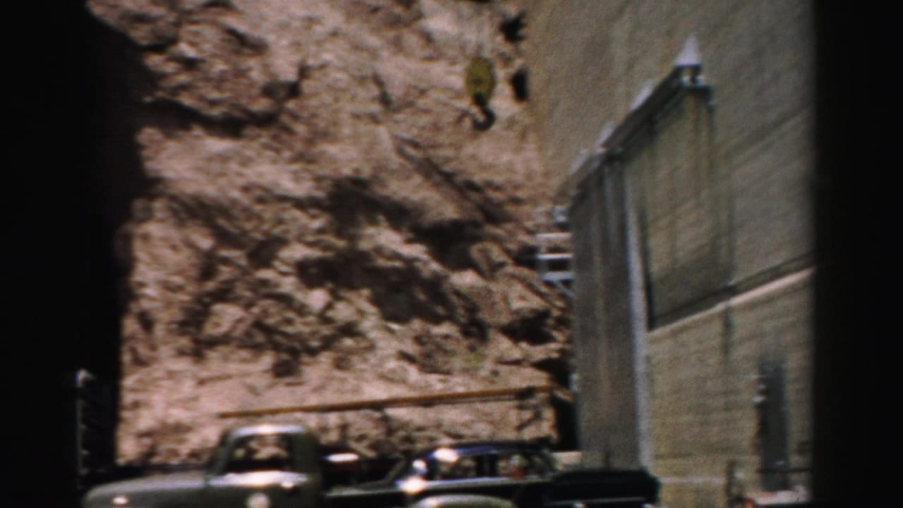 1957 Nevada 8mm frame showing a green truck near a rock face, industrial wall, railings, and parked cars.