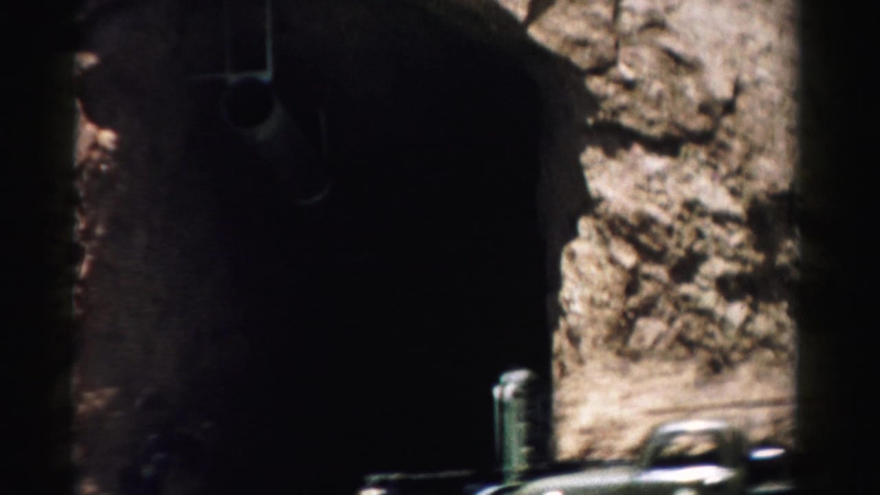 1957 Nevada home-movie frame with a shadowed tunnel opening, rough rock face, railing, and partial parked vehicles.