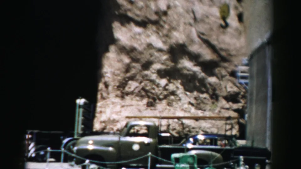 1957 Nevada frame showing the green truck below a rugged rock face, with railings, parked cars, and an industrial edge at right.