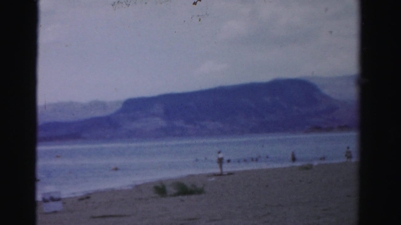 Wide sandy shoreline with calm water, distant mountains, and small figures near the water.