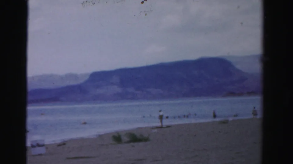 1957 Nevada 8mm shoreline view with a sandy beach, calm lake water, distant mountains, and small people near the edge.