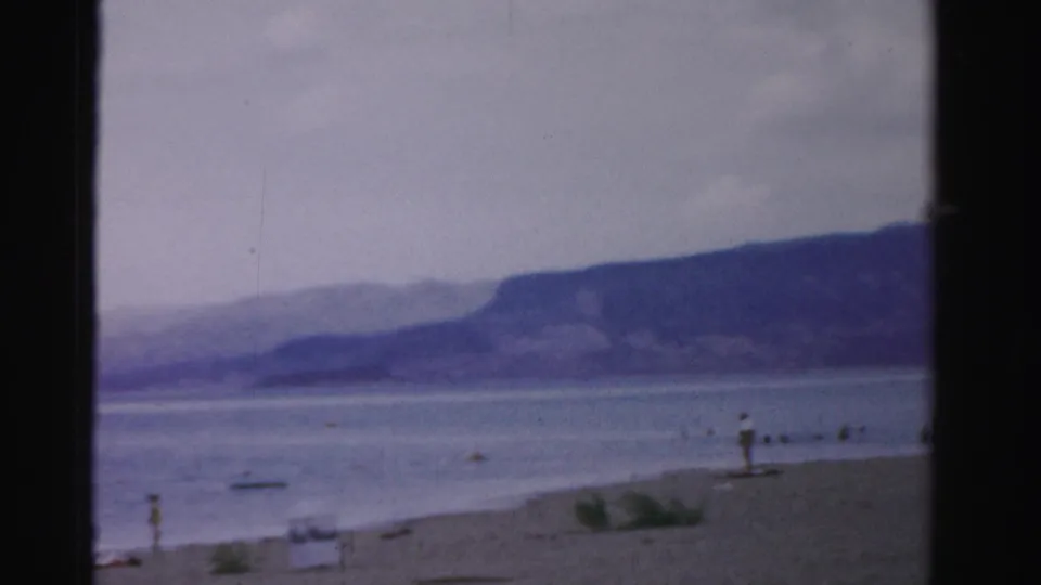 1957 Nevada home-movie frame of a sandy lakeshore with calm water, distant mountains, and small beachgoers near the shore.