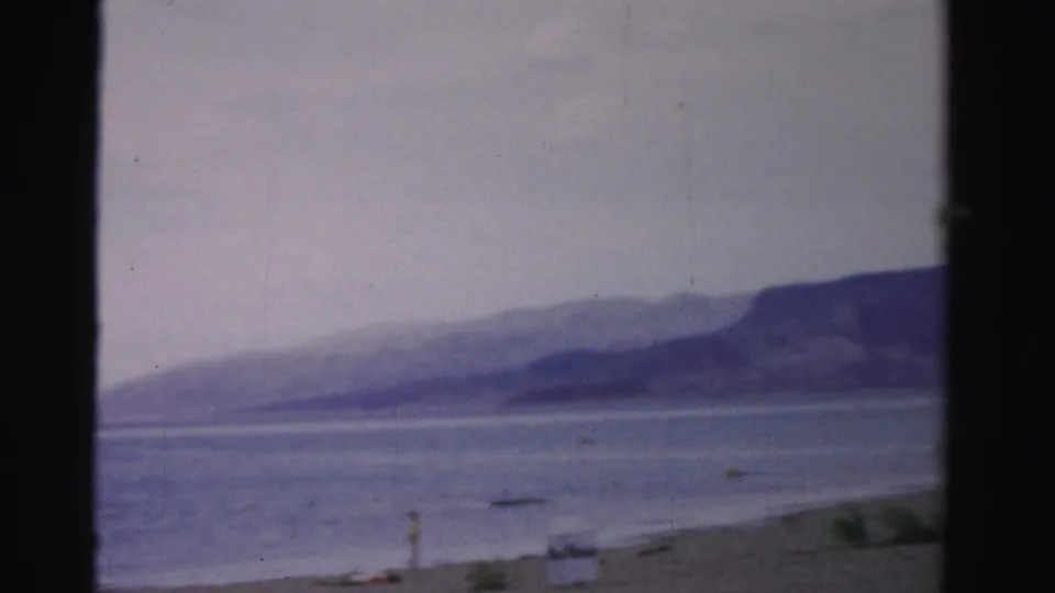 Soft 1957 Nevada 8mm frame showing a sandy shoreline, calm water, distant mountains, and a few people near the beach.