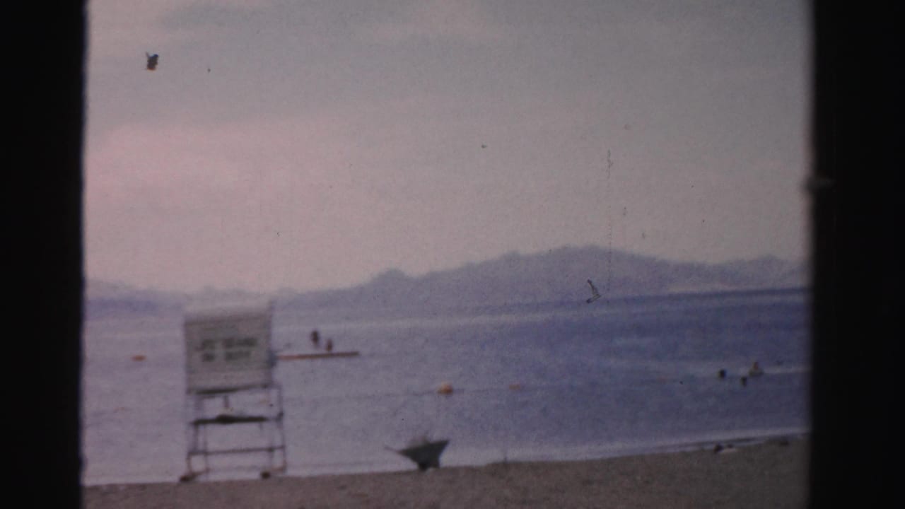1957 Nevada 8mm frame showing a sandy lakeside beach, lifeguard chair, swimmers in the water, and hazy mountains beyond.