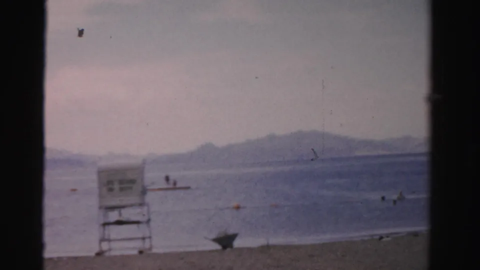 1957 Nevada 8mm frame showing a sandy lakeside beach, lifeguard chair, swimmers in the water, and hazy mountains beyond.