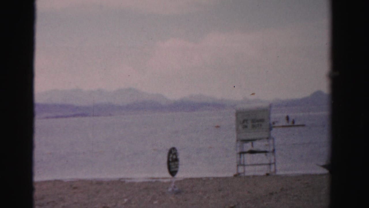 1957 Nevada lakeshore view with a lifeguard chair, small beach sign, swimmers, calm water, and hazy mountain ridges.