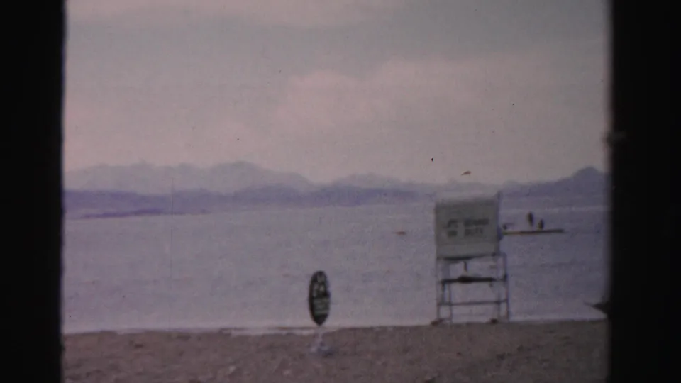 1957 Nevada lakeshore view with a lifeguard chair, small beach sign, swimmers, calm water, and hazy mountain ridges.