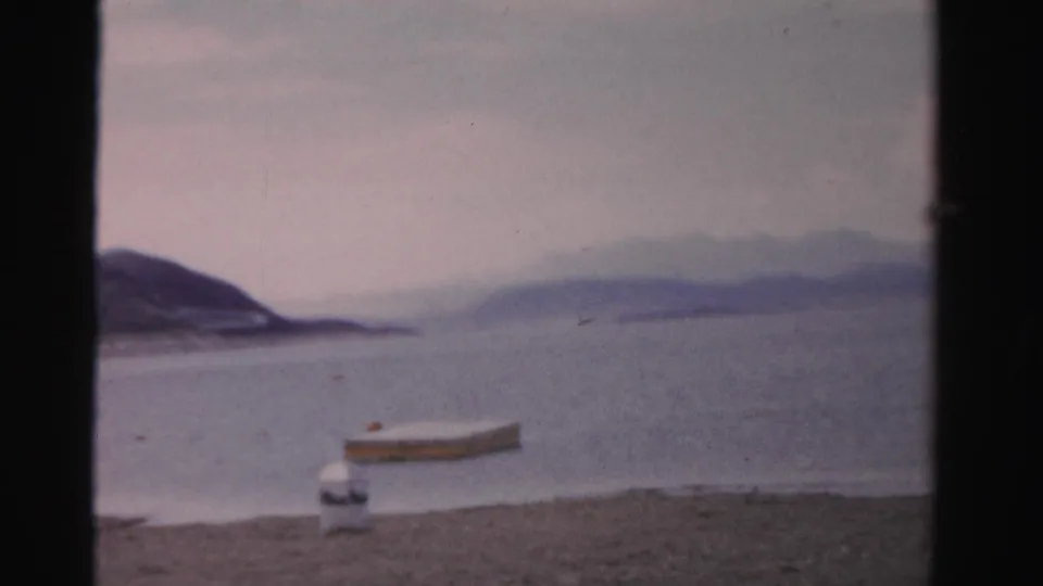 1957 Nevada 8mm view across a quiet lake from a sandy beach, with a small boat or dock shape near shore and mountains beyond.