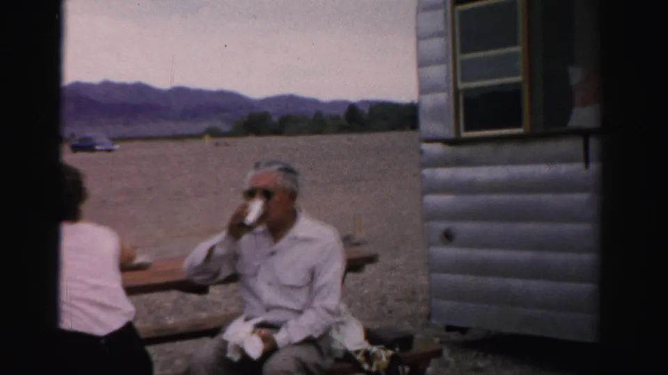 1957 Nevada lakeside home-movie frame with a man in sunglasses drinking at a picnic table beside another person near a trailer or small structure.