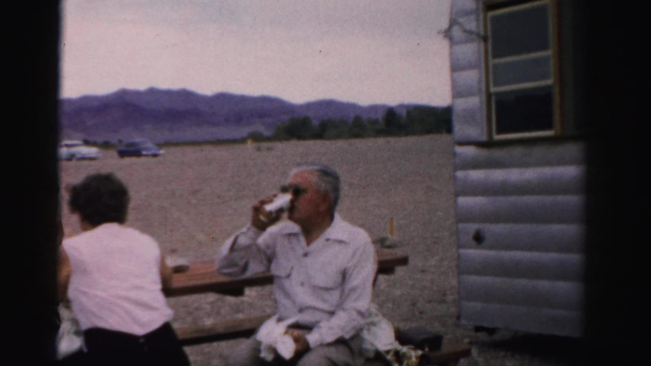 Adults seated at a picnic table beside a trailer, with one man drinking from a can.