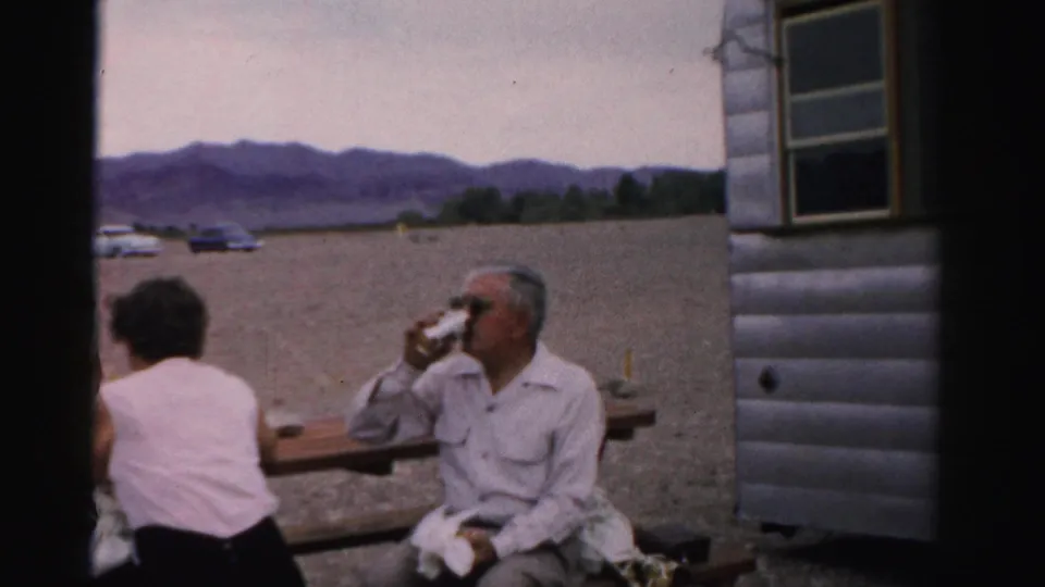 In 1957 Nevada home-movie footage, adults sit at a picnic table near a trailer while a man in sunglasses drinks from a can.
