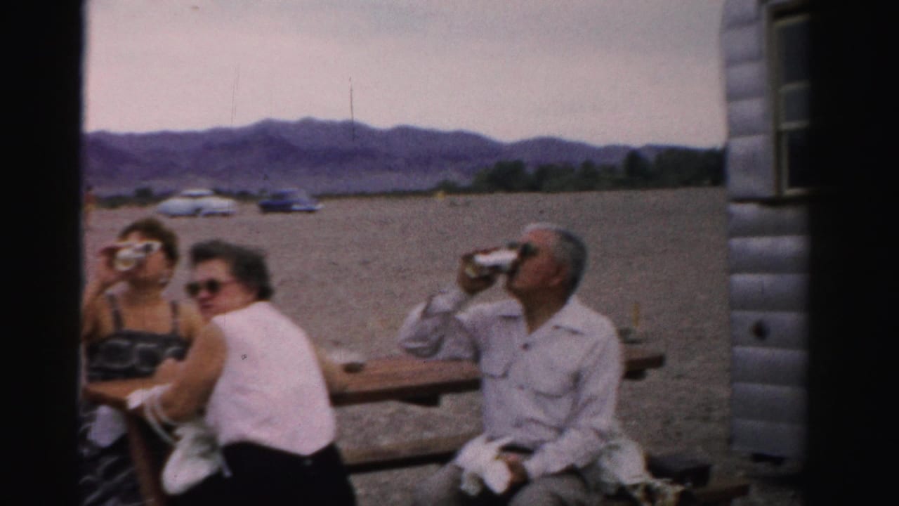 Three adults sit at a trailer-side picnic table in the supplied 1957 Nevada setting, with gravel, cars, and mountains in the background.