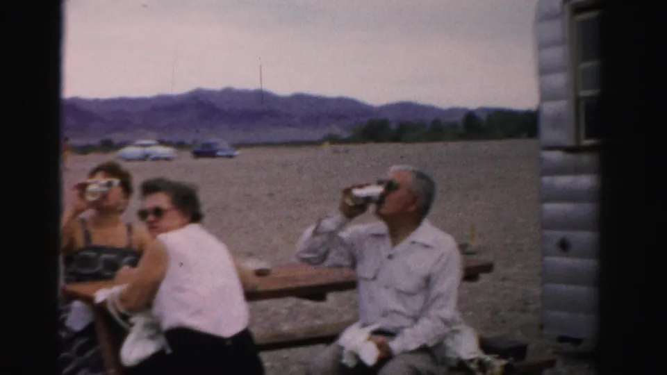 Three adults sit at a trailer-side picnic table in the supplied 1957 Nevada setting, with gravel, cars, and mountains in the background.