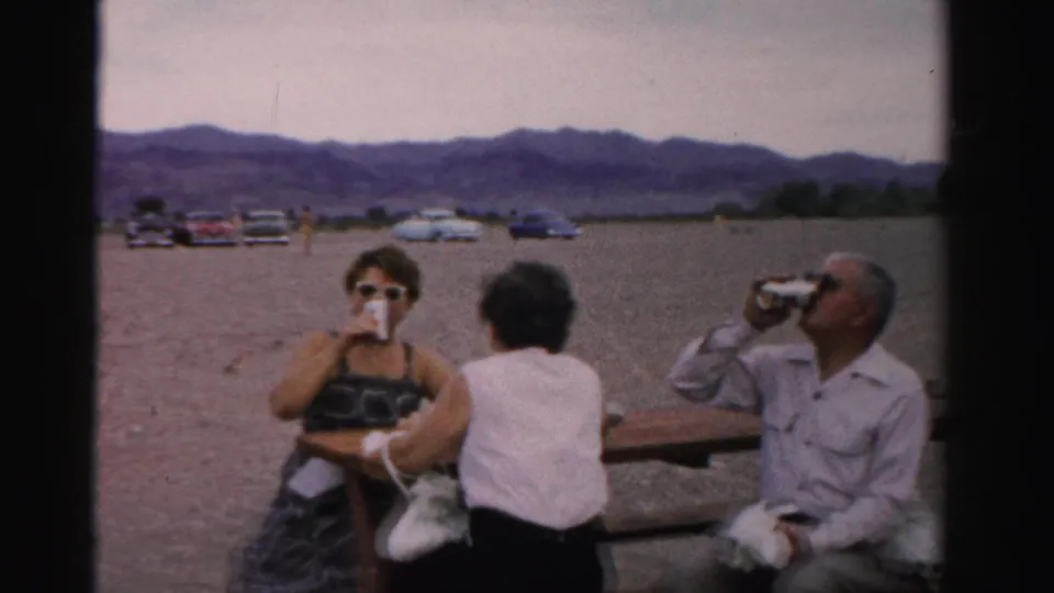 A woman in sunglasses drinks from a can while two other adults sit at a picnic table in a gravel Nevada travel setting.