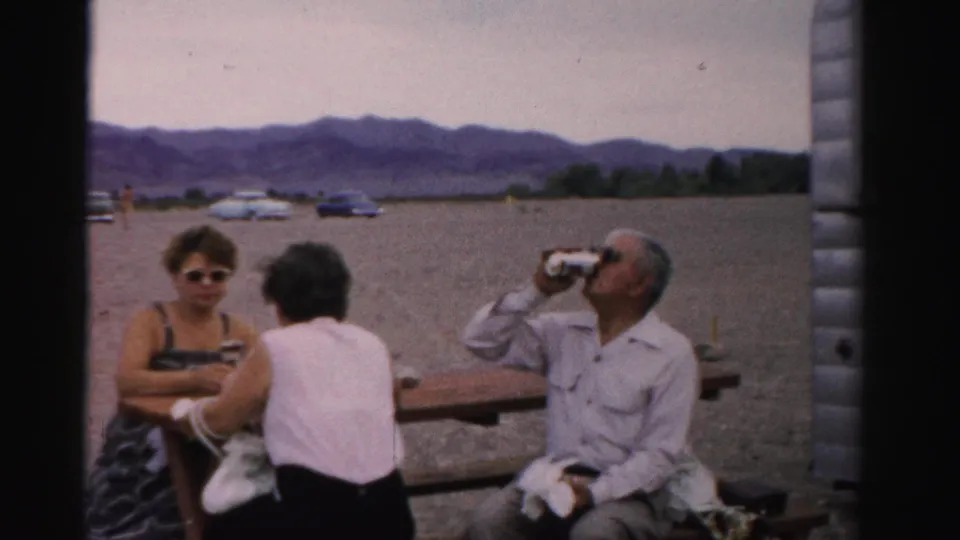 Adults remain seated at the picnic table beside a trailer as the man drinks from a can, with 1950s cars and Nevada mountains behind them.