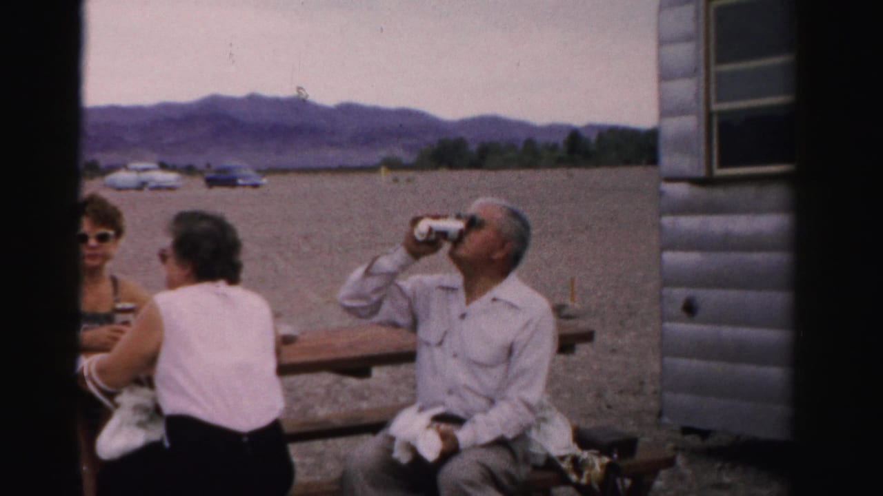 1957 Nevada home-movie frame of adults at a lakeside picnic table near a mobile home, with cars on gravel and mountains in the distance.
