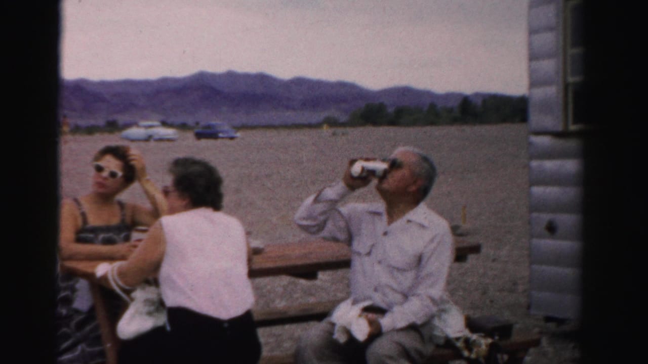 Adults at a Nevada lakeside picnic table in 1957, with one man drinking from a can and mid-century cars parked beyond the gravel area.