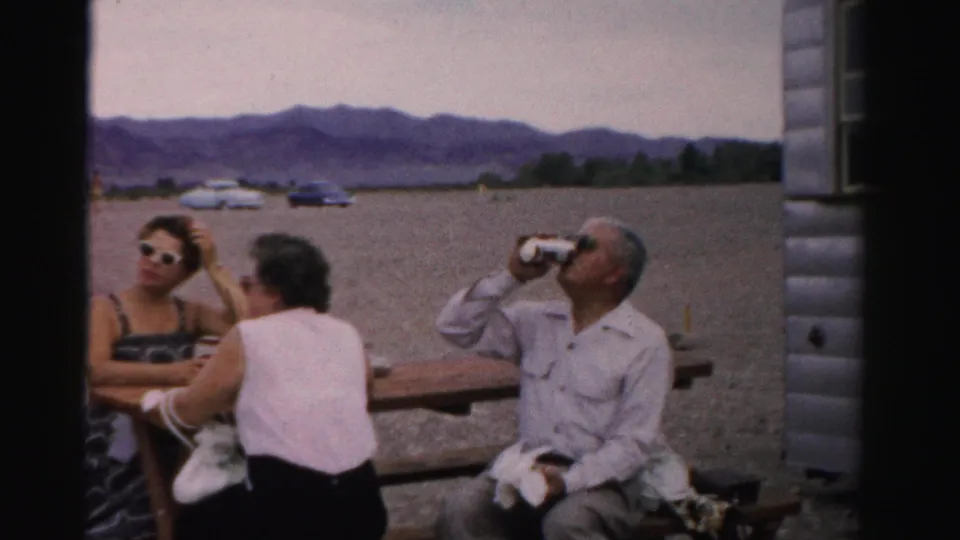 Adults at a Nevada lakeside picnic table in 1957, with one man drinking from a can and mid-century cars parked beyond the gravel area.