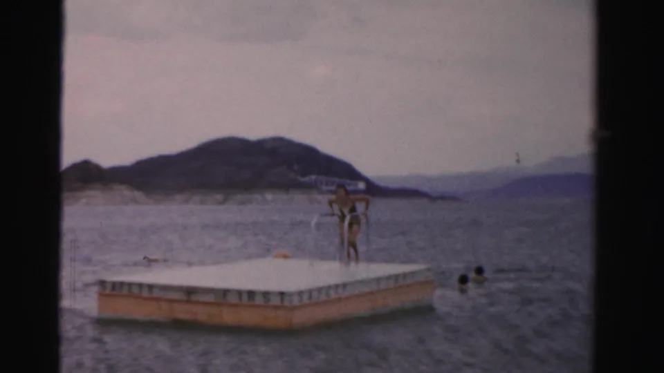 A woman in swimwear stands on a lake float marked SAFETY FIRST in this 1957 Nevada home-movie frame, with swimmers nearby and mountains beyond.