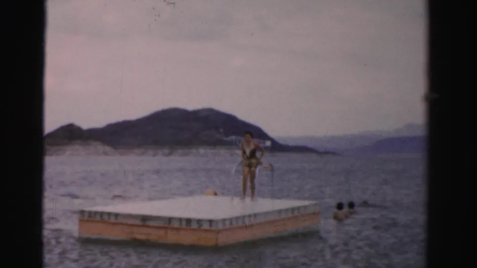 1957 Nevada home-movie frame showing a woman on a floating swim platform, with lake water, distant swimmers, and low hills in the background.