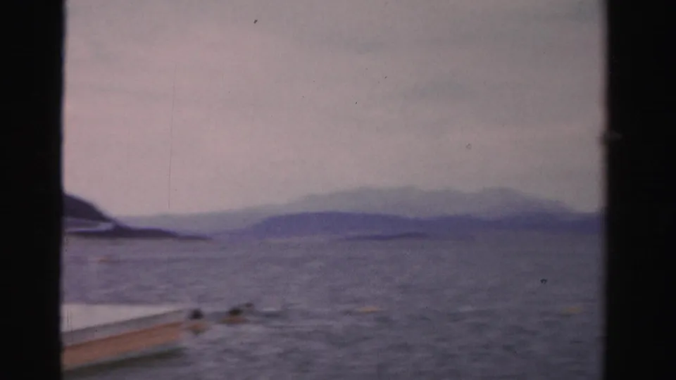 1957 Nevada home-movie frame showing swimmers beside a floating platform corner, with open water and hazy hills stretching across the background.