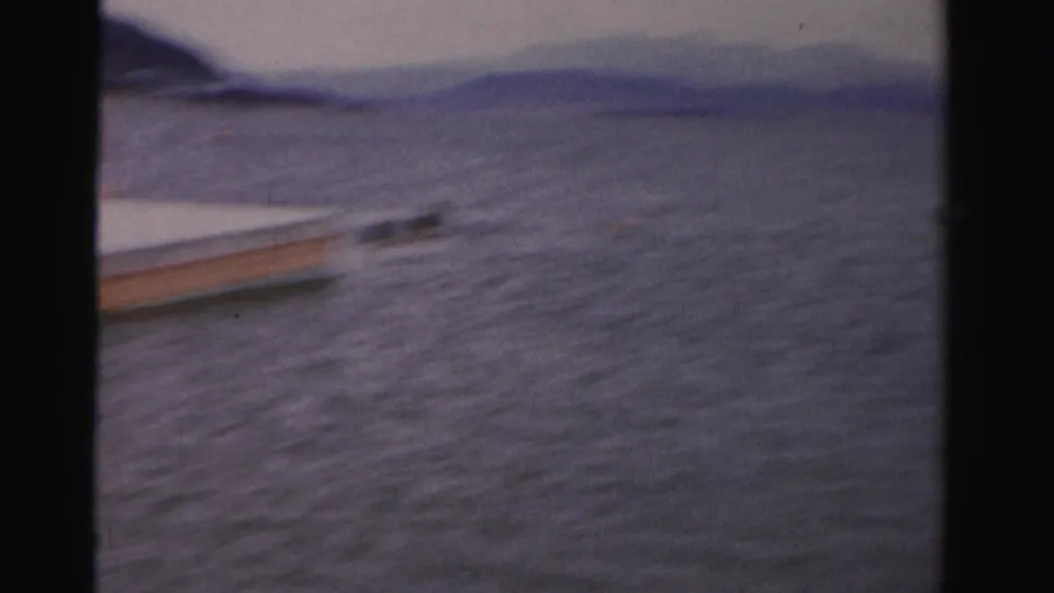 1957 Nevada home-movie frame showing the edge of a floating platform at left, rippled lake water, and low hills under a pale sky.