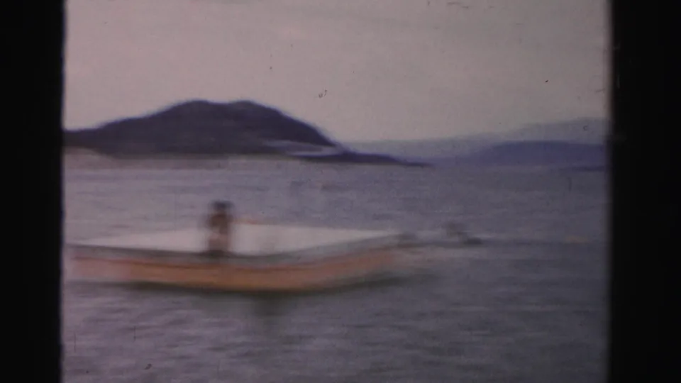 1957 Nevada 8mm frame showing a small boat or dock on calm lake water with a low mountain shoreline in the distance.