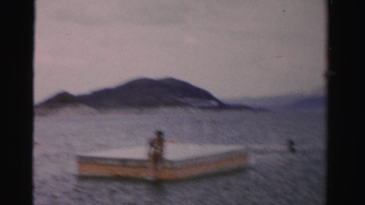 1957 Nevada home-movie frame with a person on a small boat or floating dock, calm lake water, and low mountains beyond.