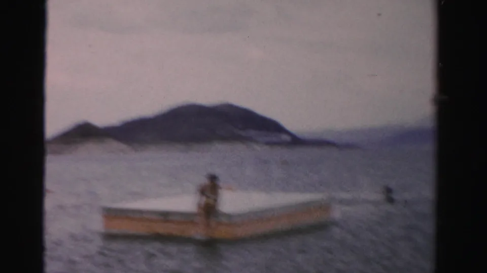 1957 Nevada home-movie frame with a person on a small boat or floating dock, calm lake water, and low mountains beyond.