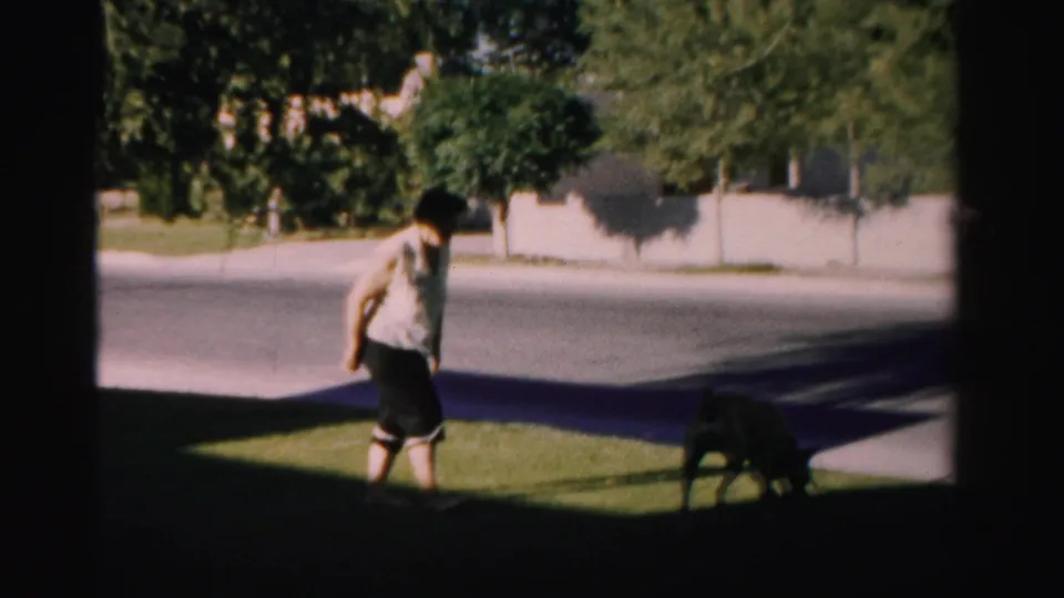 1957 Nevada 8mm yard scene with a man on a grassy lawn near a dog, pavement, trees, and a pale wall beyond.