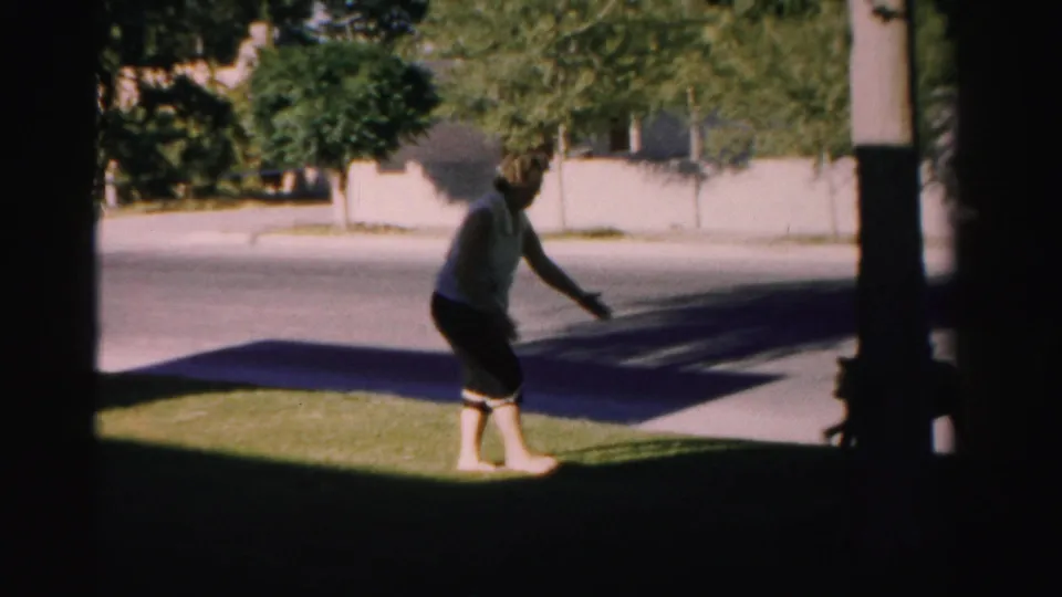 1957 Nevada home-movie frame showing a man moving on a lawn near a partly visible dog, with street, trees, and wall in the background.