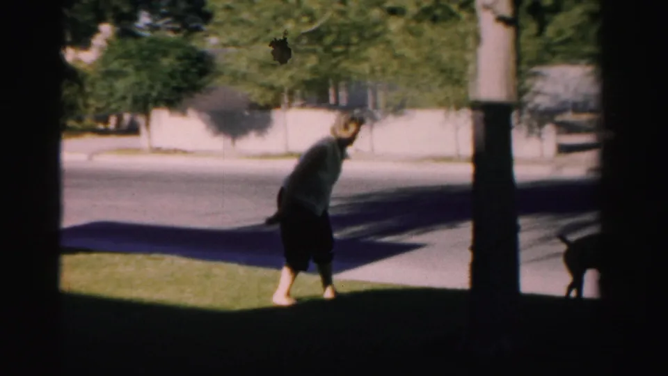 In 1957 Nevada home-movie footage, a woman bends on a shaded lawn near trees as a dark dog moves nearby.