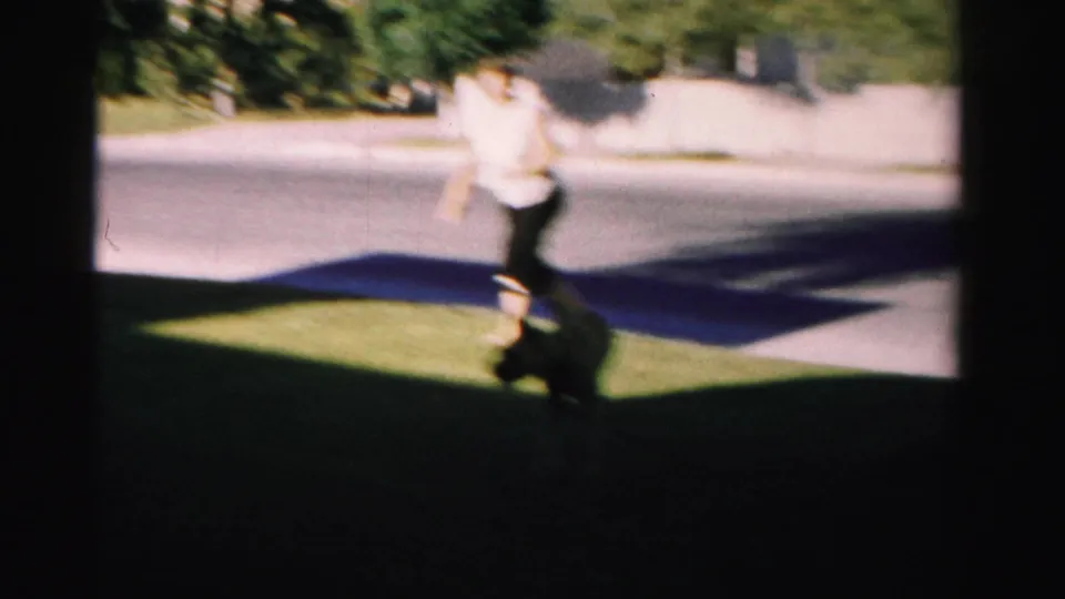 A woman runs across the shaded suburban lawn as a dark dog moves close to the grass in the foreground.