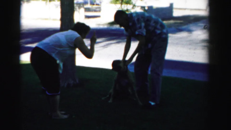 In 1957 Nevada home-movie footage, a woman and man lean toward a seated dog on grass beside a sunlit driveway.