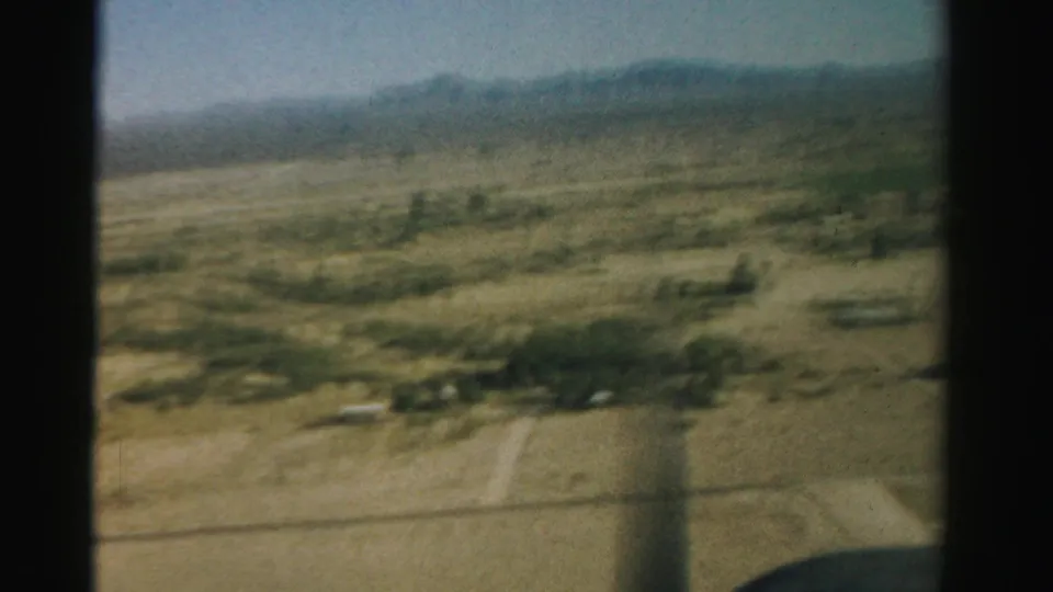 1957 Nevada aerial home-movie frame showing arid fields, scattered vegetation, mountains, and dark airplane-window edges.