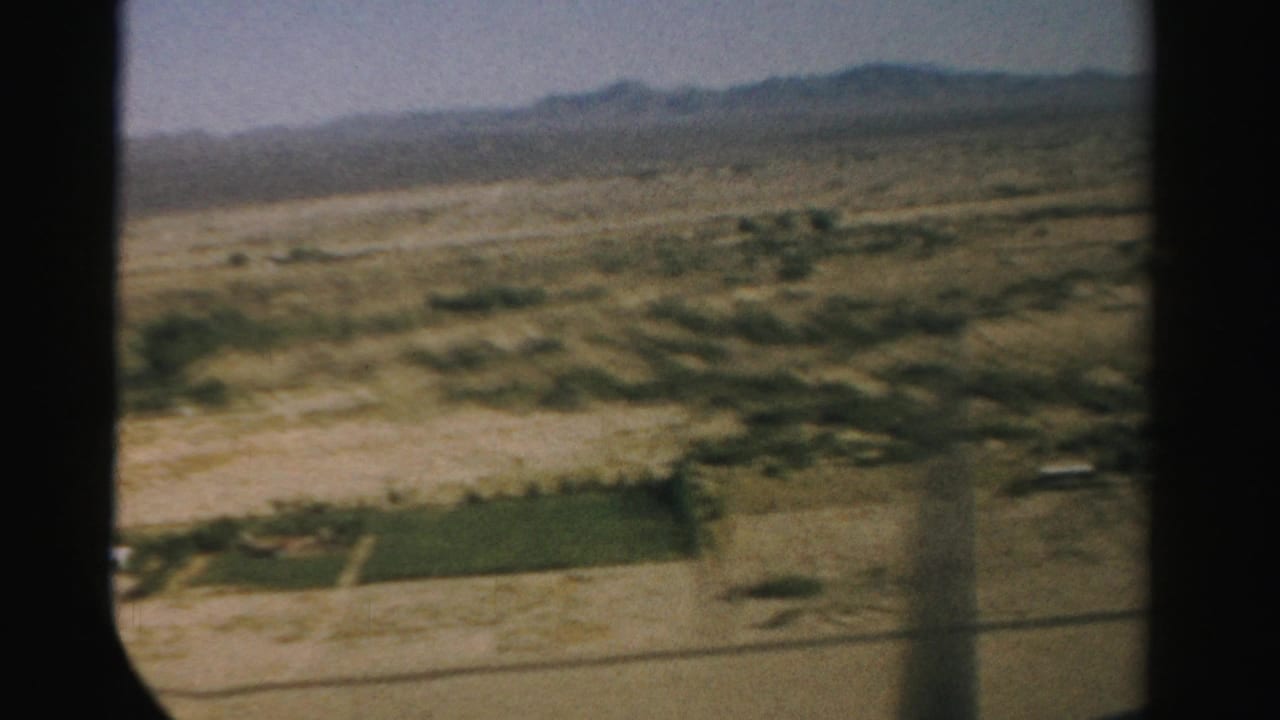 Desert farmland and mountain range seen past a dark airplane window frame.