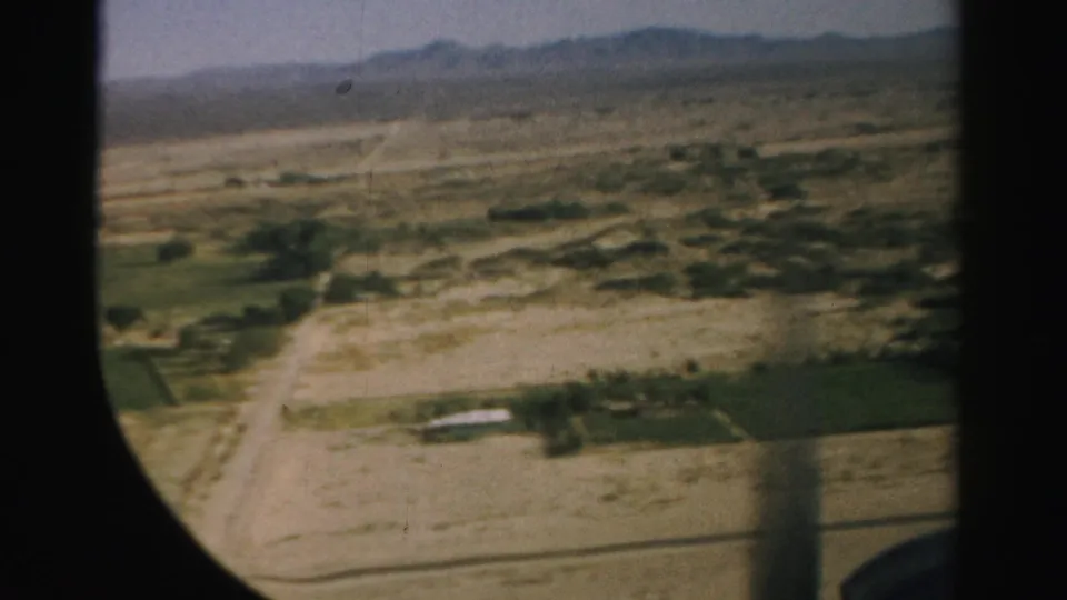 1957 Nevada aerial frame showing dirt roads, cultivated plots, dry open land, and a distant mountain ridge through the aircraft window.