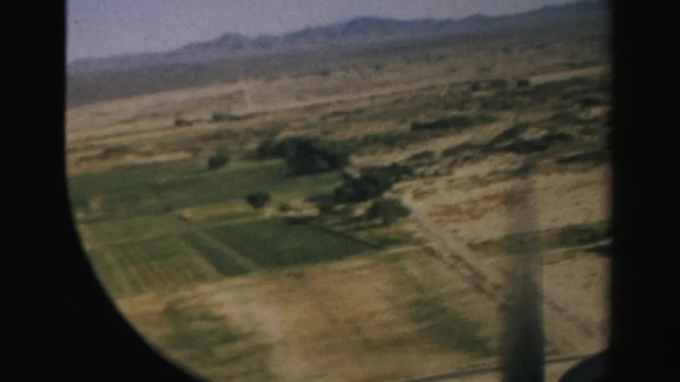1957 Nevada airplane-window view with green cultivated plots, pale desert terrain, faint roads, and mountains under a hazy sky.