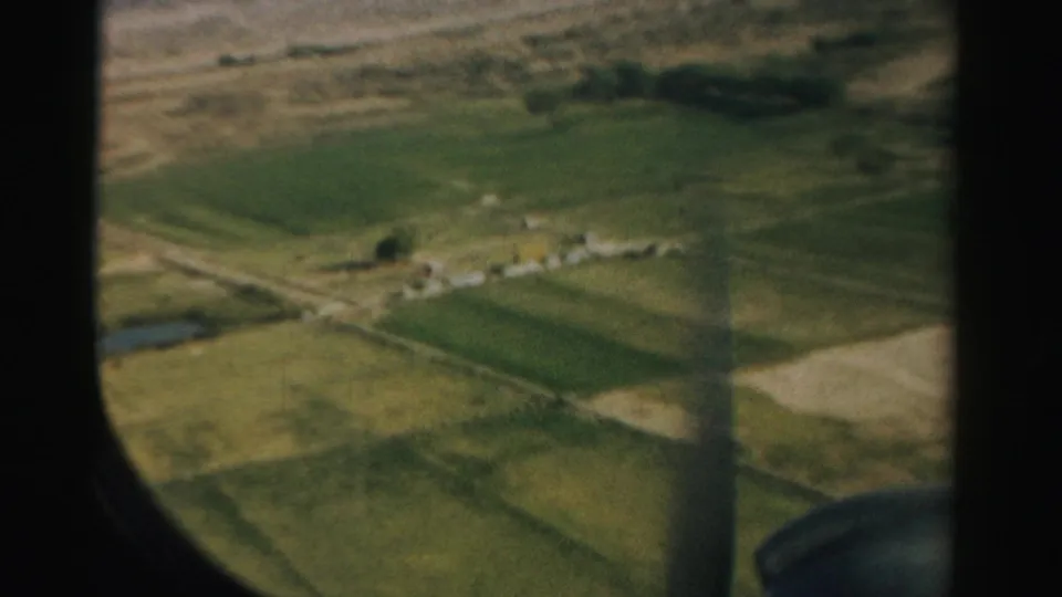 1957 Nevada 8mm frame looking through a rounded airplane window at desert terrain, cultivated fields, small structures, and a pond-like water feature below.