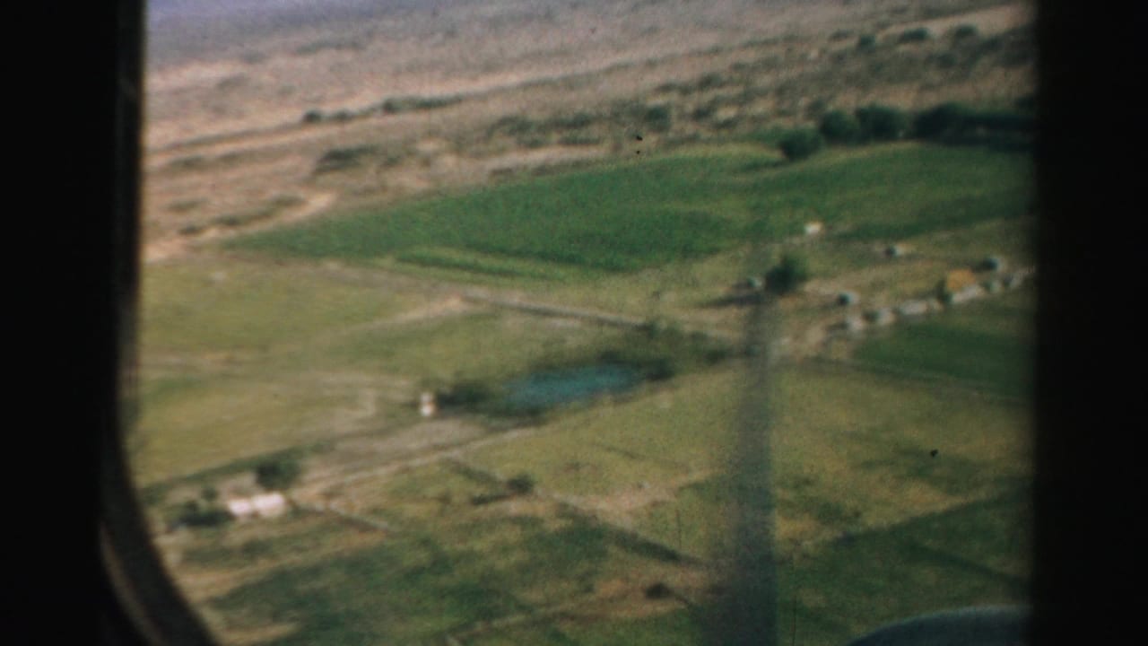 Aerial view of green fields, dry ground, small buildings, and a dark airplane-window edge.