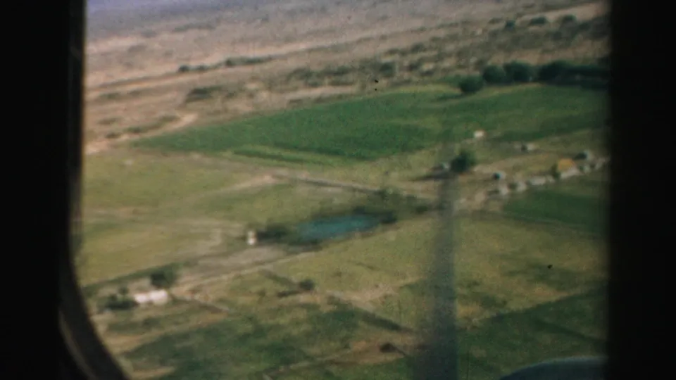 1957 Nevada aerial home-movie frame showing cultivated plots and scattered rural structures beneath the dark edge of an airplane window.