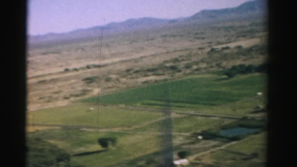 1957 Nevada 8mm frame from a descending plane, with green agricultural land in the foreground, arid ground beyond, and low mountains on the horizon.