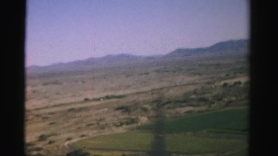 1957 Nevada home-movie frame showing dry open land and a distant mountain range from an airplane window, with cultivated fields partly visible below.
