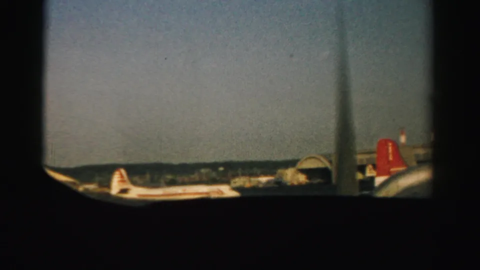 1957 Nevada airport view through an airplane window, with parked airliners, a red tail, terminal structures, and hazy sky.