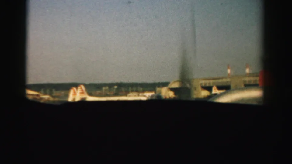 Two dark airplane window openings frame a 1957 Nevada tarmac with parked airliners and low airport structures.