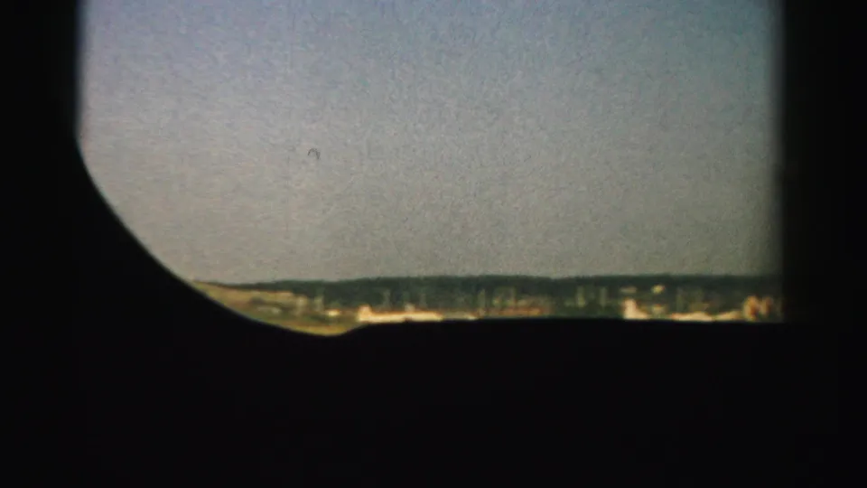 1957 Nevada 8mm frame showing a distant low landscape beneath pale sky, partly masked by a dark rounded viewfinder or window edge.