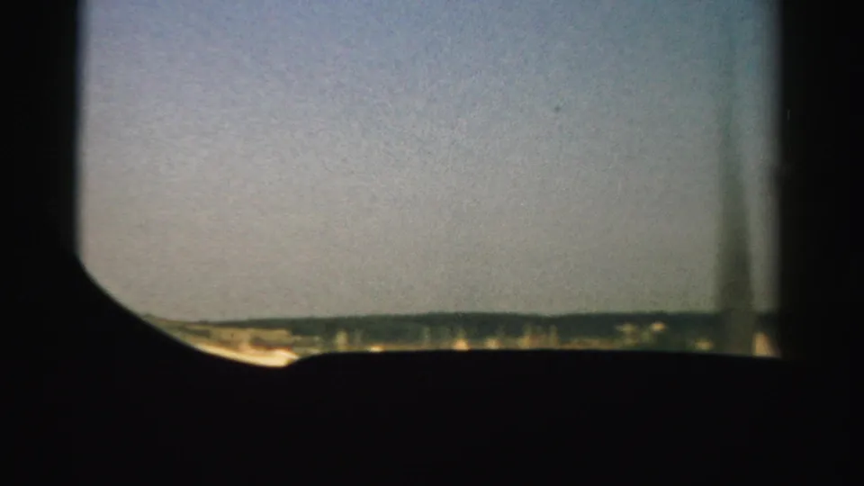 1957 Nevada home-movie frame with a distant landscape line under pale sky, seen through dark rounded viewfinder or window framing.