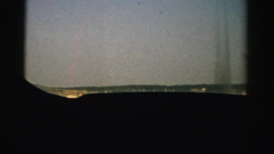 1957 Nevada home-movie frame from inside an aircraft, with a dark window edge around pale sky and runway terrain.
